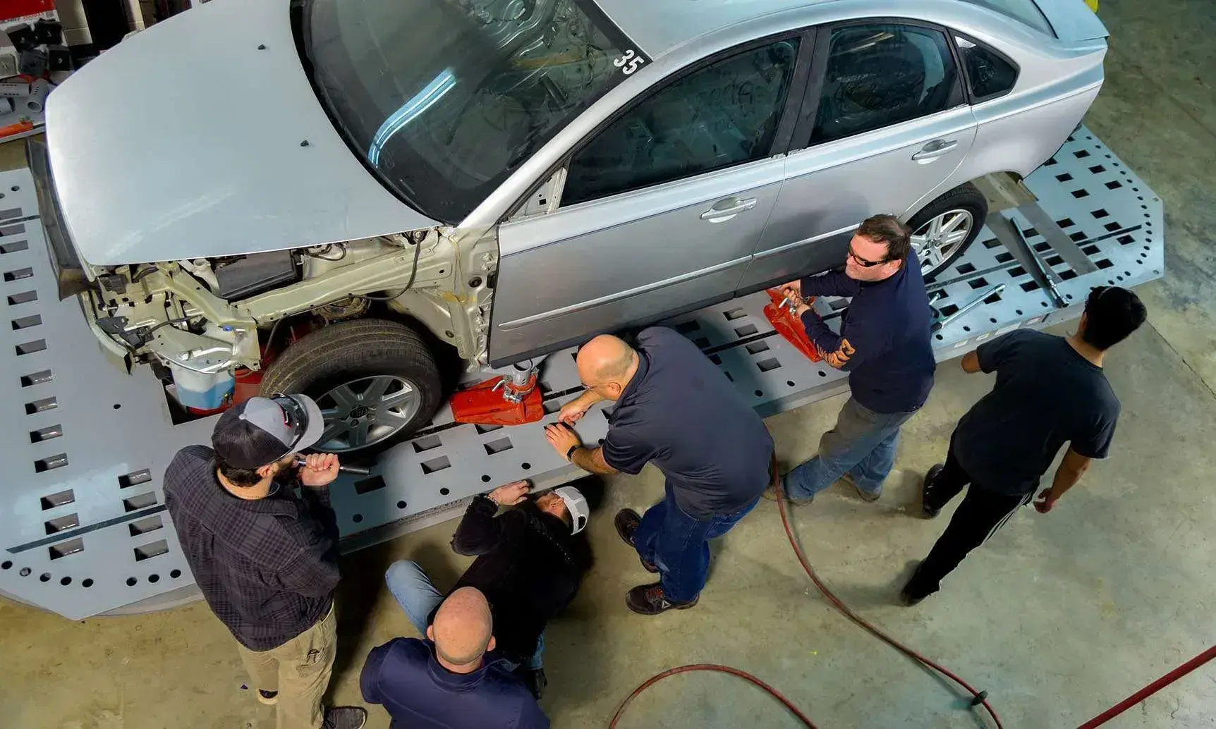 People working on a silver car in a repair shop, using tools and equipment.