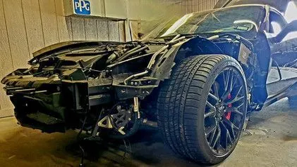 Black car with severe front-end damage, likely after a collision. The door is open, and a garage setting.