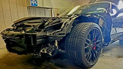 Black car with severe front-end damage, likely after a collision. The door is open, and a garage setting.