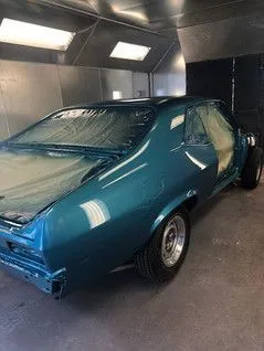 Teal classic car in a paint booth, covered in plastic, ready for finishing touches.